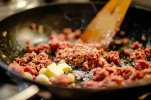 Ground beef browning in a hot skillet with a pat of melting butter, whole garlic cloves, and a wooden spatula.
