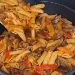 A spatula stirs a pan of cooked penne pasta, steak strips, and sliced bell peppers, all coated in a thick, savory tomato sauce.