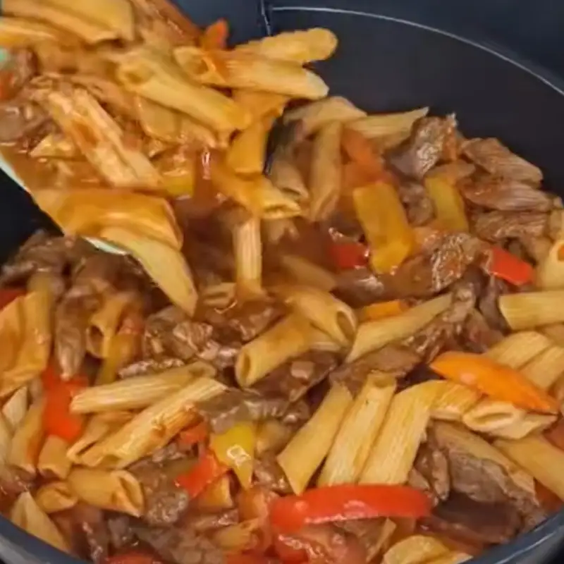 A spatula stirs a pan of cooked penne pasta, steak strips, and sliced bell peppers, all coated in a thick, savory tomato sauce.