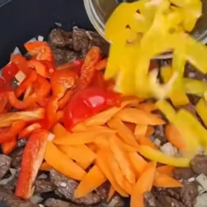 Sliced yellow bell peppers, red bell peppers, and carrots are being added to a pan containing seared steak strips and onions.