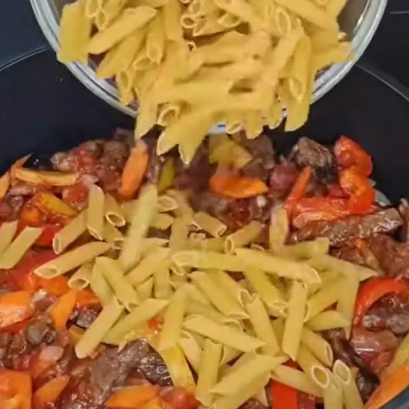 A clear bowl pours uncooked penne pasta into a black pan filled with cooked steak strips, vegetables, and tomato sauce.