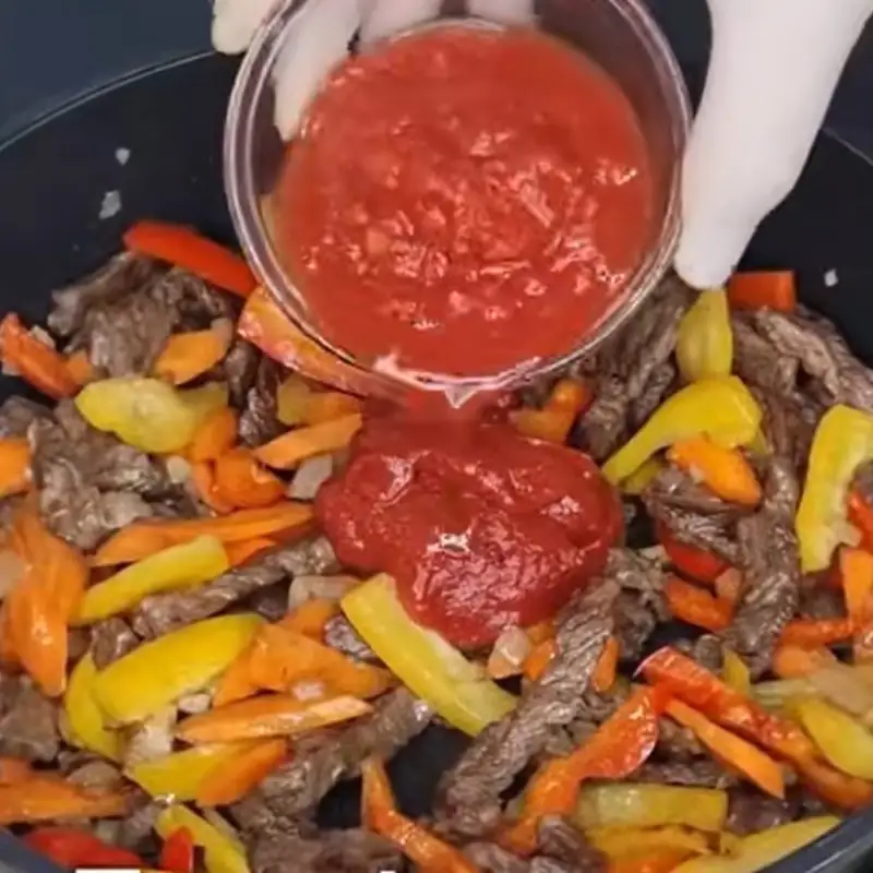 A gloved hand pours a thick red tomato sauce from a glass bowl into a pan of cooked steak strips and sautéed vegetables.