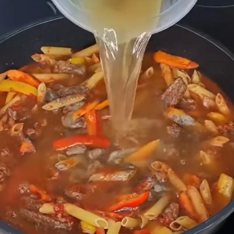  A light-colored liquid broth is being poured from a measuring cup into a pan, submerging the uncooked pasta, steak, and vegetables.