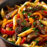 A close-up of a cast-iron skillet filled with zesty steak pasta, showing cooked penne, steak pieces, and bell peppers, garnished with fresh parsley.
