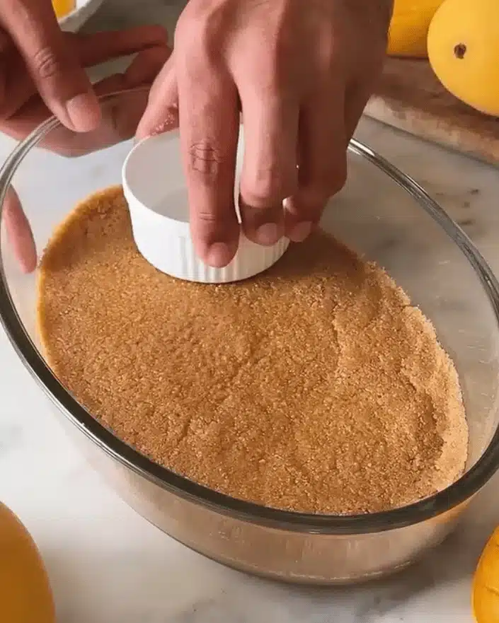 Hands using a small white bowl to press down a buttery biscuit crumb mixture into an oval glass dish to form the base for a no bake mango dessert.
