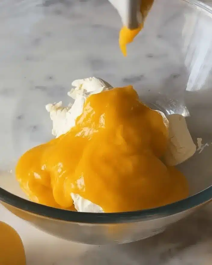 Vibrant mango puree being added to white cream cheese in a clear glass bowl, beginning the mixture for a no bake mango dessert filling.