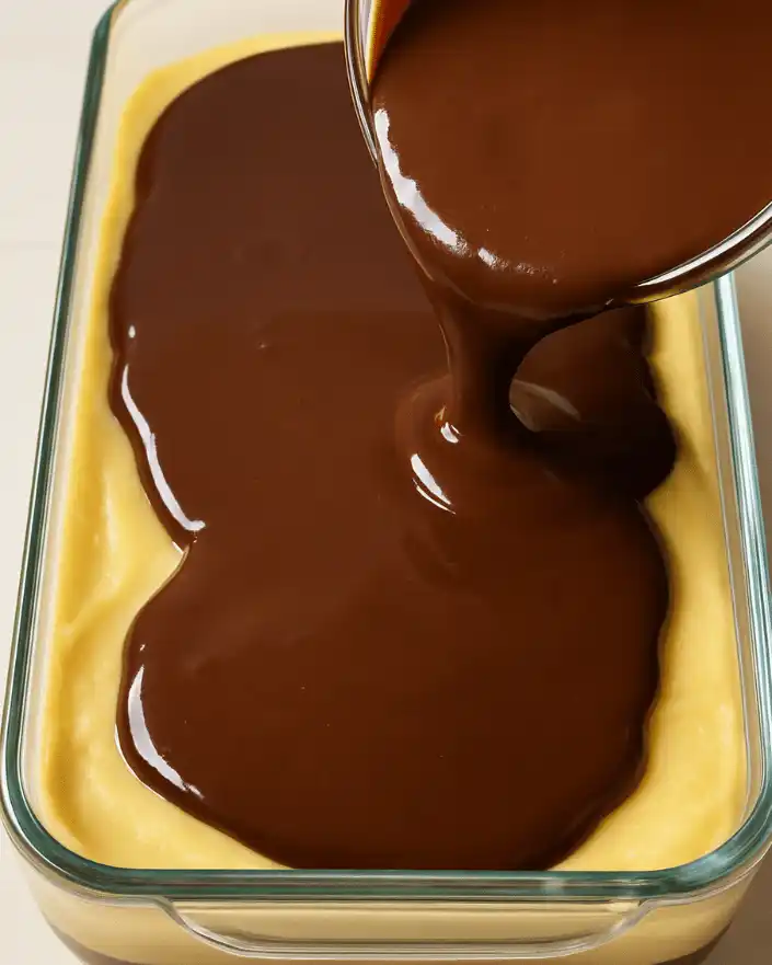 Chocolate ganache being poured over custard layer in glass dish for no bake Boston cream pie bars.