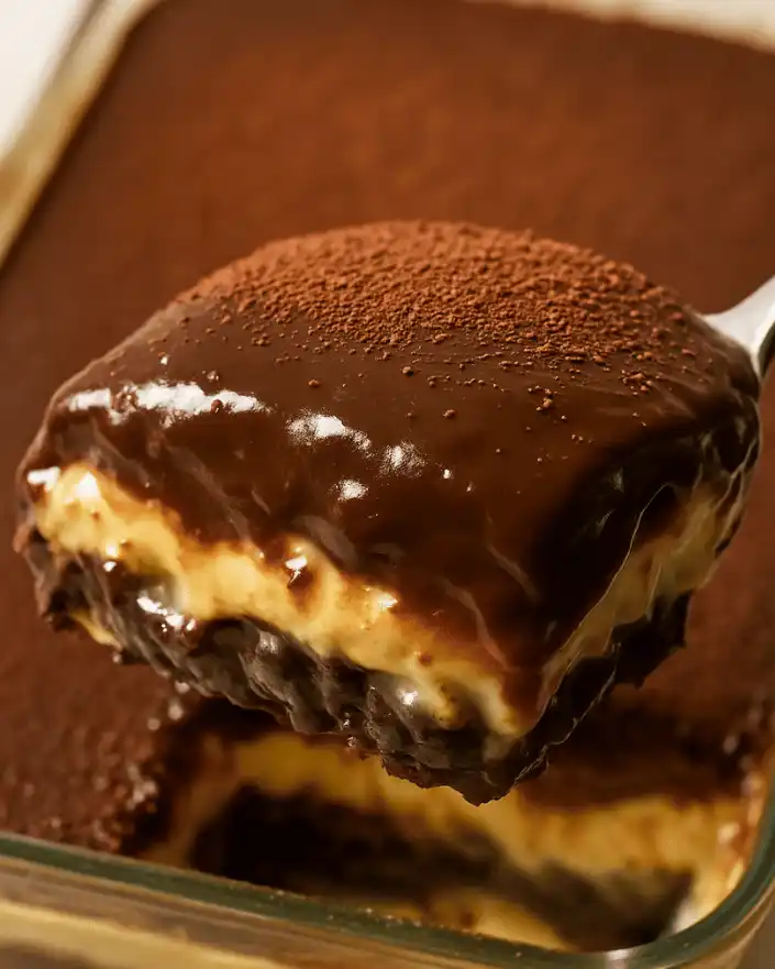 A spoon lifting a slice of no bake Boston cream pie bar showing biscuit, custard, and chocolate ganache layers.