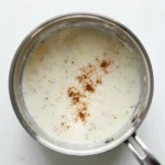 Creamy Bechamel Sauce 2 Close-up overhead photo showing freshly grated nutmeg sprinkled over the surface of the finished Creamy Bechamel Sauce in a saucepan for final seasoning.