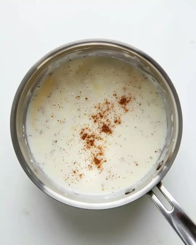 Creamy Bechamel Sauce 5 Close-up overhead photo showing freshly grated nutmeg sprinkled over the surface of the finished Creamy Bechamel Sauce in a saucepan for final seasoning.