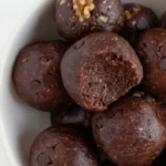 No-Bake Brownie Bites 2 Close-up of fudgy no-bake brownie bites in a white bowl, showing a bite taken out of one to reveal a rich texture with visible chocolate chips, walnuts, and almonds.